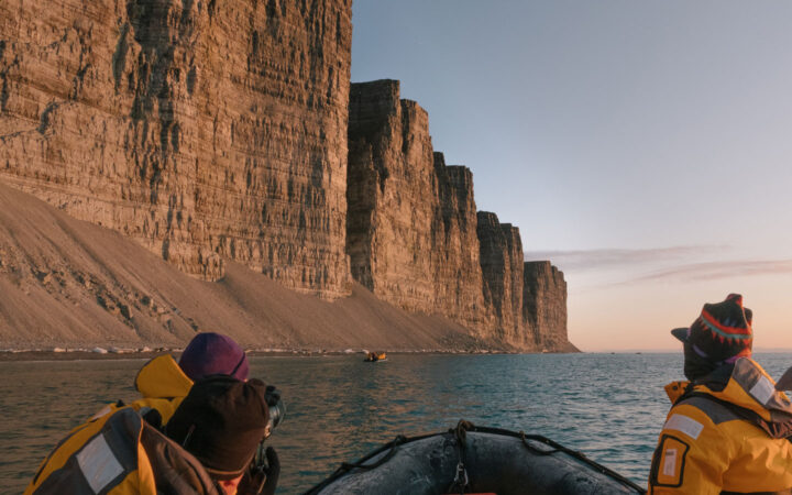 3 Northwest Passage cruise guests in yellow coats ride in a Zodiac boat along tall tan cliffs at sunset.