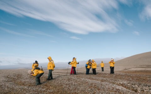 Small group of Northwest Passage cruise guests in yellow jackets walk over brown tundra & take photos on a sunny day.