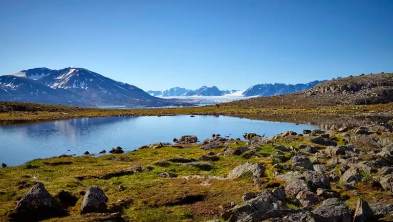 Glassy, small glacial lake surrounded by rocky green tundra & snowy peaks on a clear day during a Svalbard In Depth cruise.
