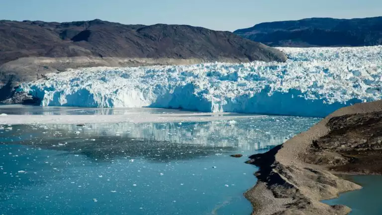 Eqi glacier of compressed snow & ice sits beside glassy glacial water with brown hills on either side, seen in the Thule region.