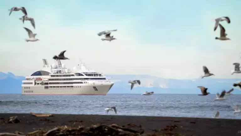 Small ship with gray & red hull cruises past dark shoreline as seagulls fly above during a cruise to Greenland & Baffin Bay.