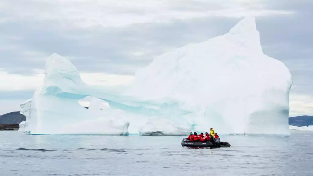 Thule region travelers in red parkas ride a Zodiac boat beside a massive iceberg in calm water under cloudy skies.