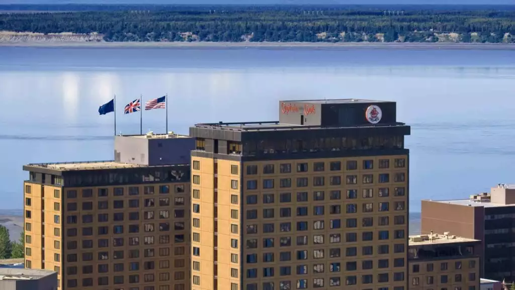 Aerial view of top of Captain Cook Hotel in Anchorage, with 3 beige towers overlooking calm sea with green mountains behind.