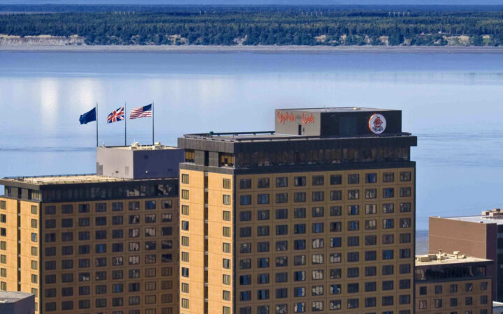 Aerial view of top of Captain Cook Hotel in Anchorage, with 3 beige towers overlooking calm sea with green mountains behind.