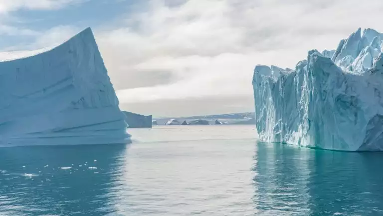 2 large icebergs sit in calm glacial water under cloudy sky, seen on an In the Ice of the Arctic, Greenland to Svalbard cruise.