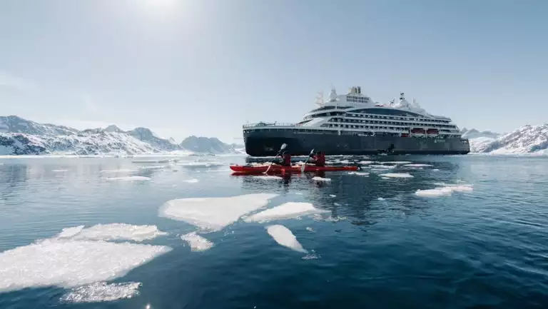 Le Commandant Charcot small ship with blue hull & white upper decks sits in sunshine while tandem kayakers paddle red boats by ice.