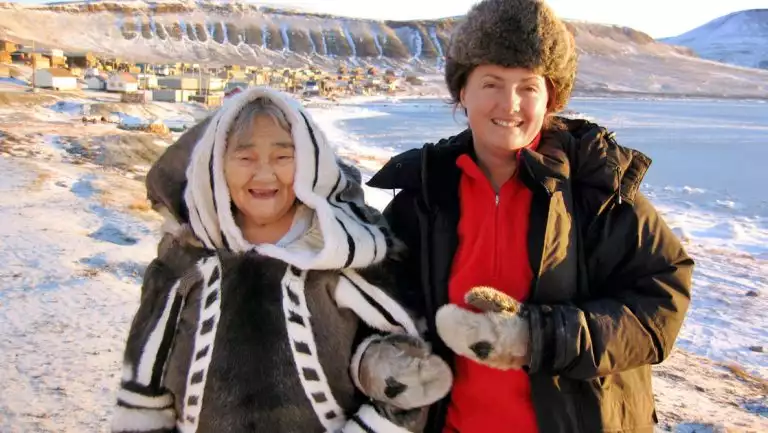 Older woman in warm Inuit garb stands beside younger woman in brown jacket & furry hat on a sunny day in the Thule region.