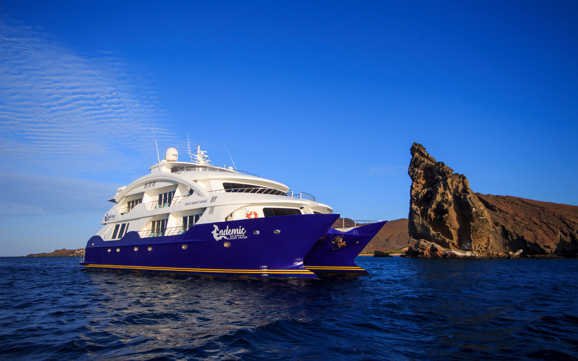 Golden Galapagos catamaran with blue twin hulls & 2 white upper decks sits in calm water by a large brown rock on a sunny day.