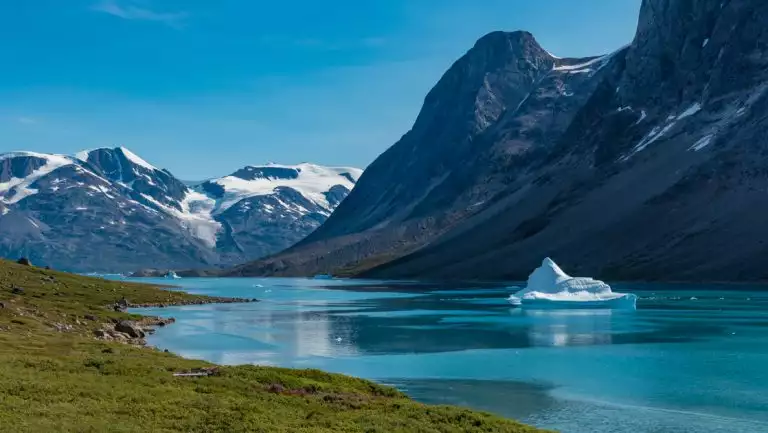 Iceberg sits in blue water on a windy day beside green shores & dark, rocky mountains partly covered in snow, seen in Greenland.