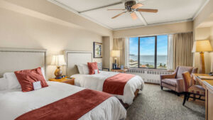 Guest room at Hotel Captain Cook in Anchorage with 2 queen beds in white linens with red accents, chair, desk & view window.