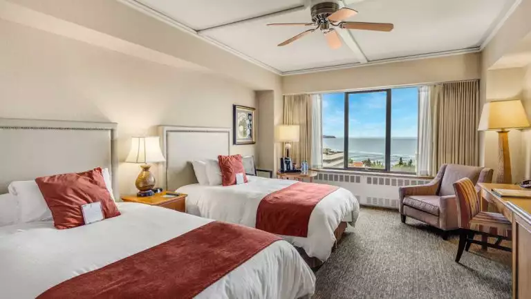 Guest room at Hotel Captain Cook in Anchorage with 2 queen beds in white linens with red accents, chair, desk & view window.