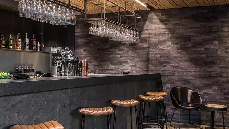 Empty stone bar at Vik, Iceland's Hotel Kria, with leather & metal stools, hanging wine glasses, wood ceiling & soft lighting.
