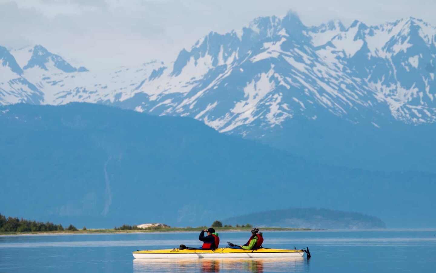 Two Alaska travlers kayaking in a calm bay with large jagged mountains in the background in Alaska.