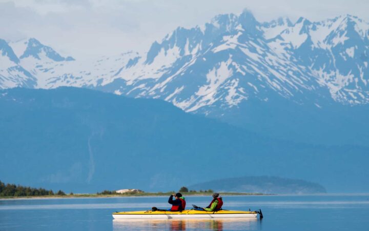 Two Alaska travlers kayaking in a calm bay with large jagged mountains in the background in Alaska.