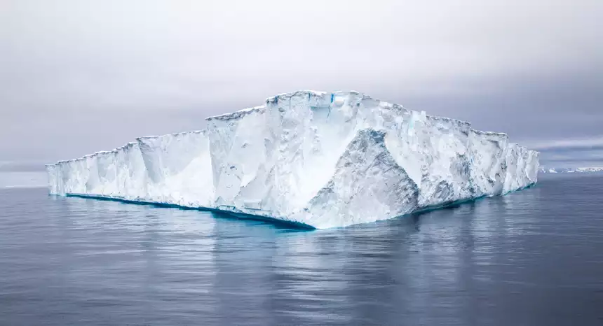 A massive white flat topped tabular iceberg the size of a house floats in the dark blue ocean of Antarctica in November.
