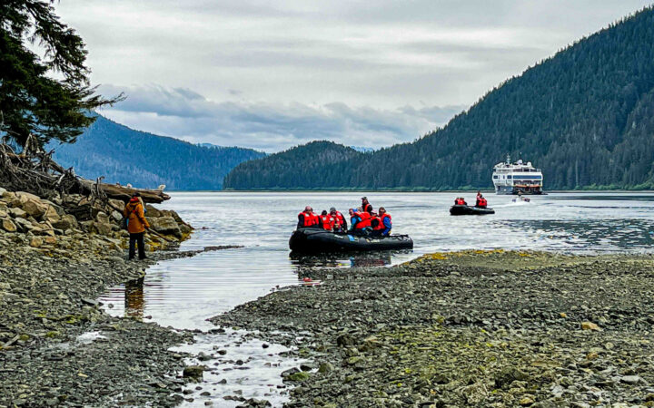 2 Zodiac boats with Alaska travelers pull into pebble shoreline with guide standing in tan coat on a Lindblad Alaska cruise.