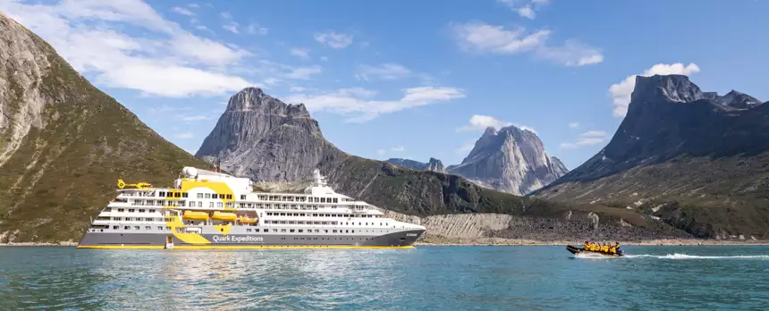 The expedition ship Ultramarine cruising on calm green water with a zodiac racing towards the bow and dramatic mountain peaks in the background.