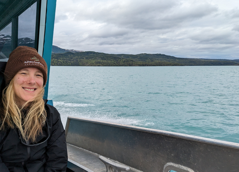 Woman in black jacket & brown knit hat sits at front of a boat in Alaska under cloudy skies.
