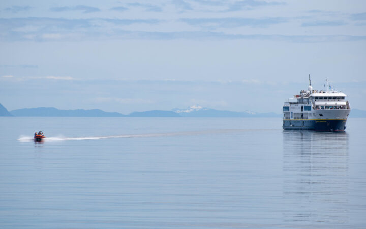Small blue & white expedition ship sits in glassy water as an orange Zodiac dinghy motors away during a Lindblad cruise.