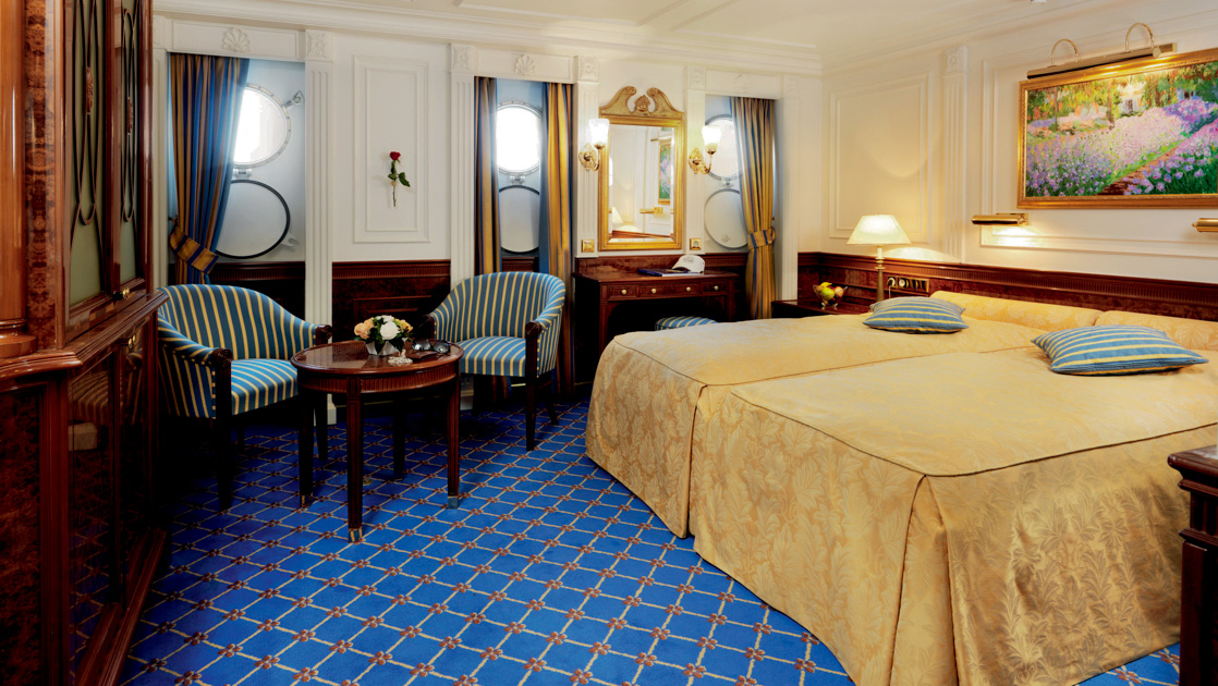 Category 3 cabin on Sea Cloud II: Lindblad with double bed in gold, blue carpet, 3 portholes, desk, 2 chairs & wood coffee table.