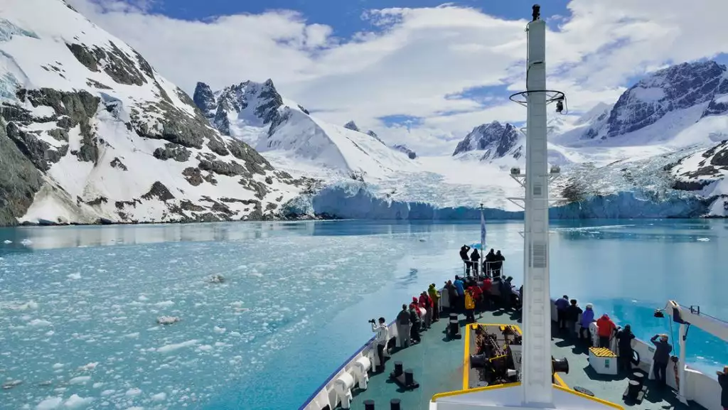 Travelers stand on bow of small ship as it cruises turquoise water with iceberg bits in the sun, toward snowy mountains.