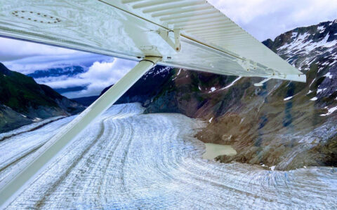 View from under white wing of small flightseeing plane, looking onto large glacier between mountains, seen in Glacier Bay.
