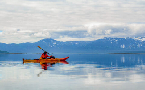 Lone kayaker in red & yellow boat paddles glassy water near tall mountains under cloudy sky on a Glacier Bay multiday tour.