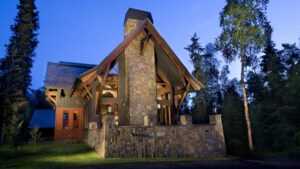 Exterior of 1 of the Homes at Kenai Riverside Lodge in Alaska with big stone patio & chimney, lawn, red & gray wood accents.