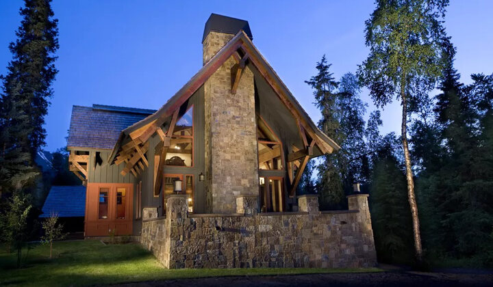 Exterior of 1 of the Homes at Kenai Riverside Lodge in Alaska with big stone patio & chimney, lawn, red & gray wood accents.
