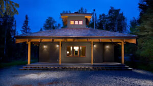 Small 1-level cottage at dusk with covered porch, 1 large window & exterior accent lights, surrounded by forest in Alaska.