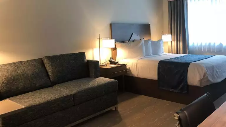 Guest room at Kodiak Compass Suites with gray couch, double bed in white & blue linens, desk with leather chair & bedside tables.