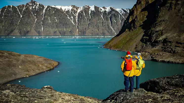 2 Ultimate Arctic Voyage travelers in yellow coats stand on cliff overlooking turquoise fjord with snowcapped mountains behind.