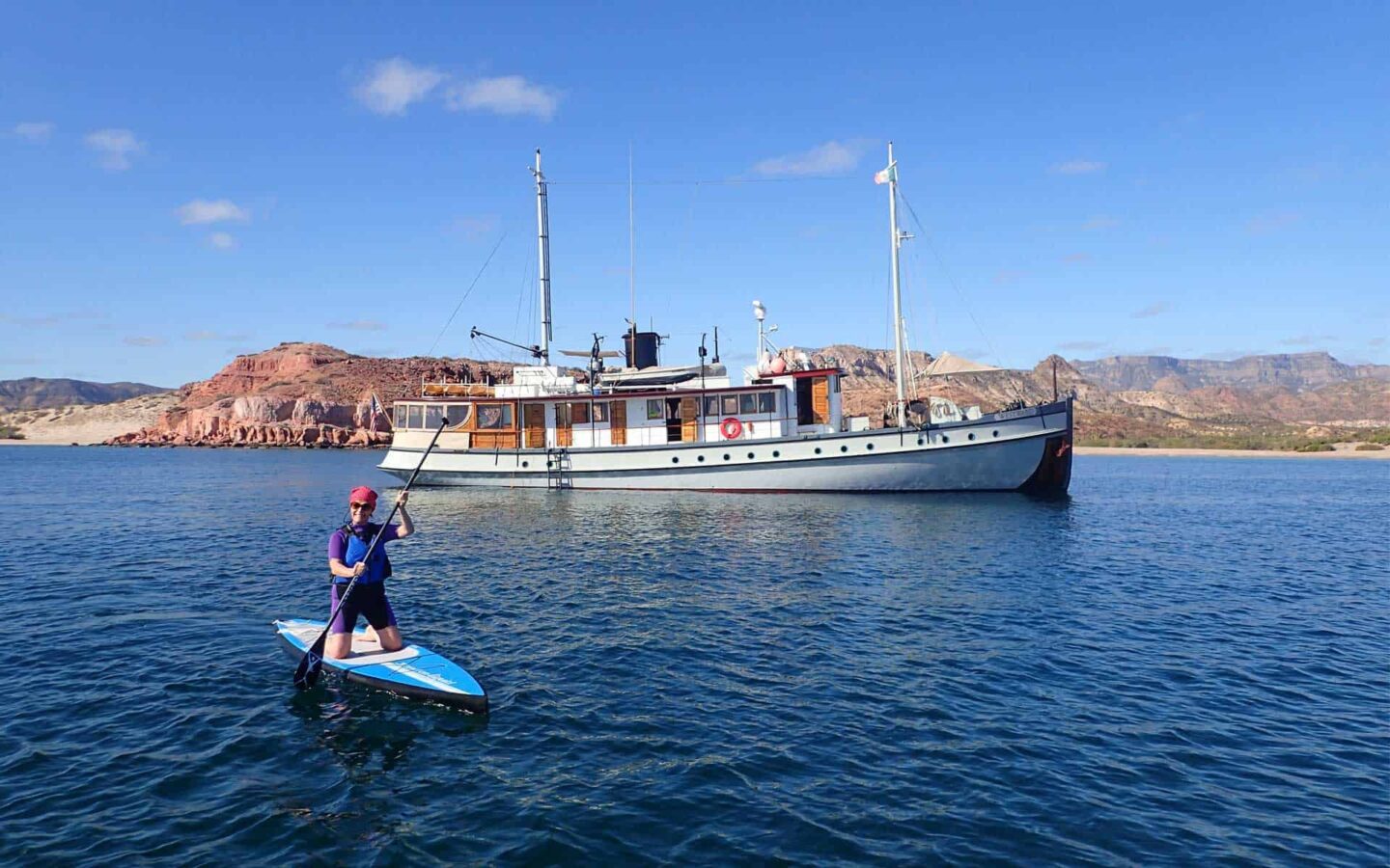 Woman paddleboarding in Baja witha small ship behind her and desert mountains.