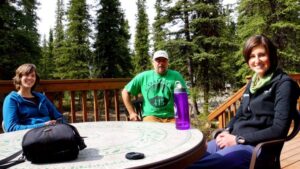 Three people sit around a table on a wooden porch surrounded by trees at Denali Education Center near Denali National Park in Alaska