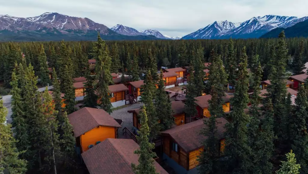 Aerial view of Denali Cabins: small wooden guest cabins among trees near the national park with mountains in the distance.