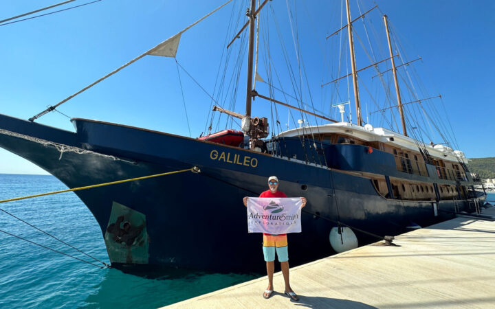 Man holds flag in front of dark blue Galileo sailing ship docked in Greece on a sunny day.