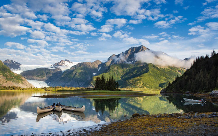 Three canoes float off shore of a lake that runs to a green mountain range and icy white and blue glacier in the distance.