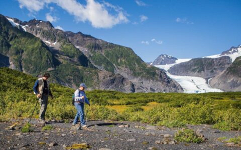 2 men hike over flat terrain with small rocks with a glacier, rocky mountain peaks & green shrubs behind in Kenai Fjords NP.
