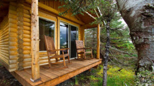 Log cabin covered porch with wooden rocking chairs, big window & sliding glass door at Kenai Fjords Glacier Lodge in Alaska.