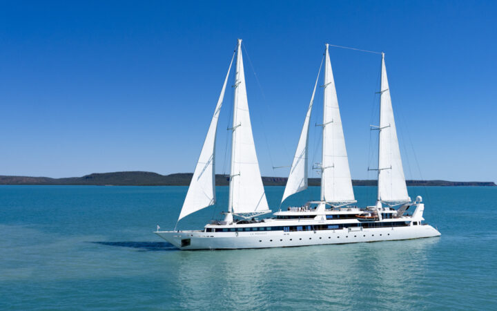 Exterior of Le Ponant white small sailing ship with 3 masts & 5 sails cruising in calm turquoise water on a sunny day.