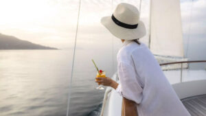 Woman in long white billowy shirt & Panama hat leans against railing & holds tropical cocktail on Sun Deck of Le Ponant ship.
