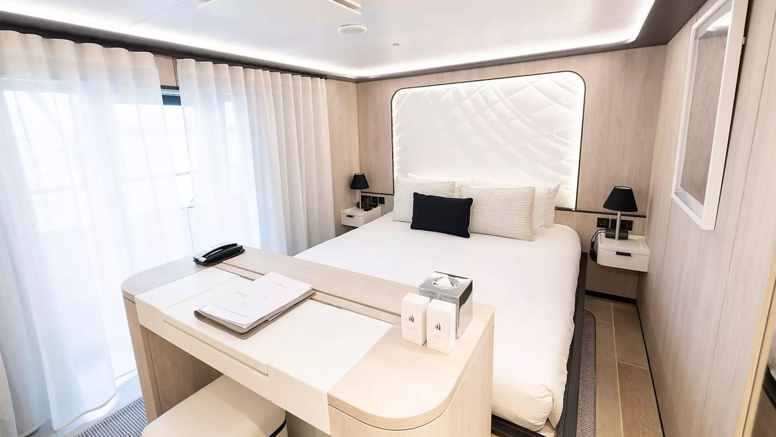 King bed in white with accent pillows & white padded headboard in beige room with glass windows to balcony on Le Ponant ship.