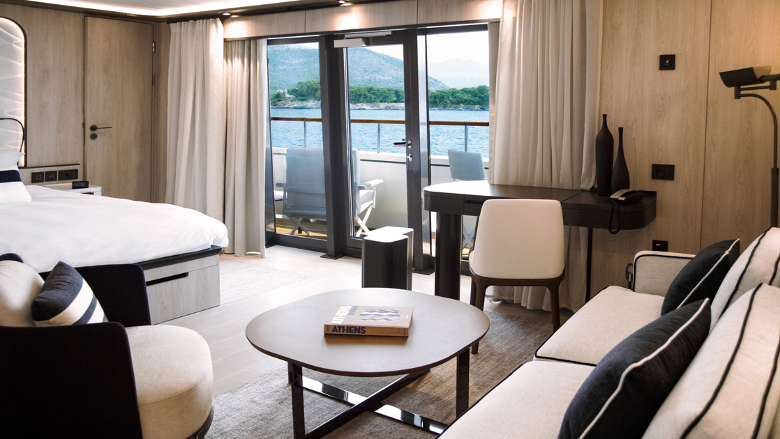 King bed with white linens & padded headboard in beige room with couch, chairs, deck & glass windows to balcony on Le Ponant ship.