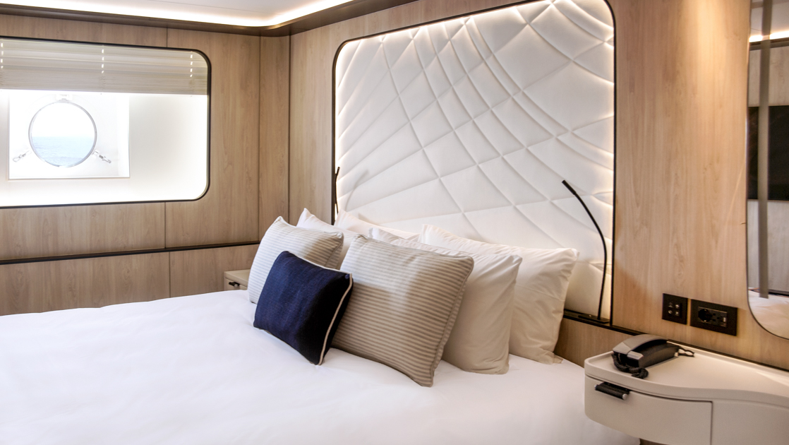 King bed in white with accent pillows & white padded headboard in beige room with porthole on Le Ponant cruise ship.
