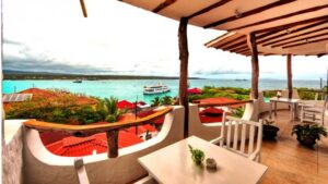 a table and chairs sitting on the balcony of the Angermeyer Waterfront Inn galapagos hotel with the ocean and a small ship in the background