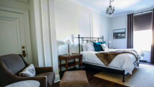 Guest room with double bed, wooden side tables, chandelier, velour chair & ottoman, wooden floors & marble table at Fort Garry Hotel in Churchill, Canada.