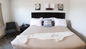 Guest room with double bed enclosed in a tan bedspread, white pillows & fleece beluga whale-shaped throw blanket, with bedside table and chair beside it at the Polar Inn in Churchill, Canada.