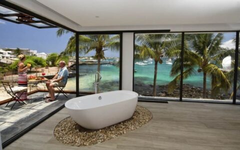 standalone bathtub on wood floor of a luxury galapagos hotel with large windows behind it overlooking the ocean and travelers sitting outside