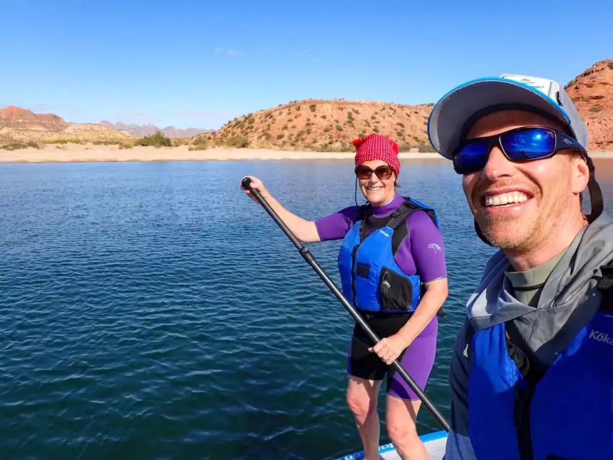 Man & woman in blue life jackets & sunglasses stand on paddleboards in calm sea near sandy beach & arid hills on a sunny day in Baja.
