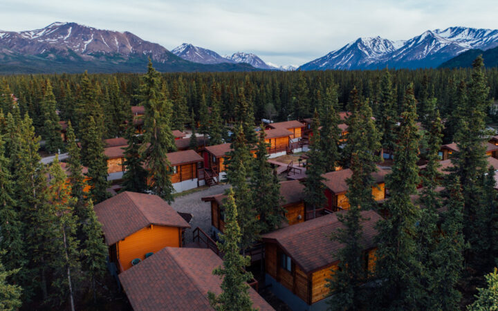 Aerial view of Denali Cabins: small wooden guest cabins among trees near the national park with mountains in the distance.
